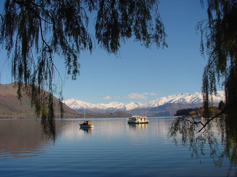 A photo of a lake with two boats in the mid-ground, a small one and a larger one, and snow-covered mountains in the background.