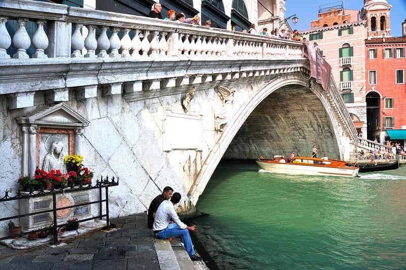 Two men sitting beside the water on stone steps, next to an arched bridge, with a cobbled area behind them, and some kind of sculpture of altar against the bridge.