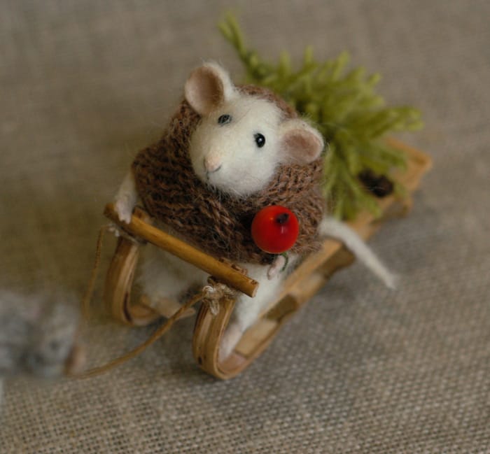 A craft work depicting a mouse with a scarf made of brown string, and a red fruit in its left hand, riding a sled with a Christmas tree in the back.