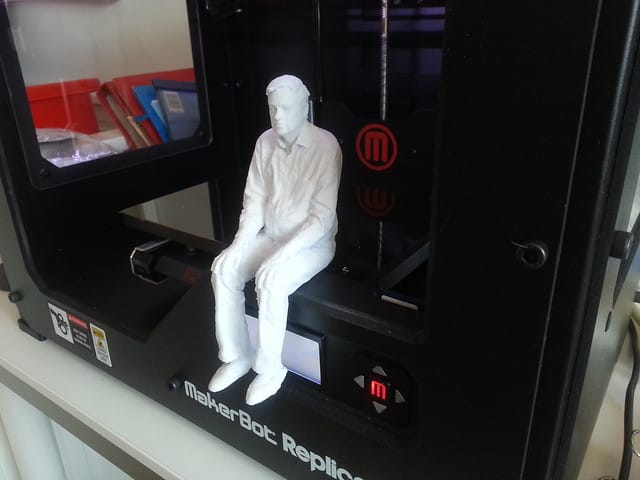 A white, 3D-printed human figure, sitting on the printing platform of a MakerBot Replicant 3D printer.