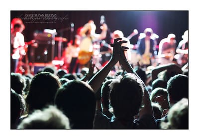 View of a music concert from within the crowd, the band in soft focus in the distance, a punter clapping his hands above his head.