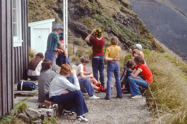 A group of young people in casual dress, sitting outside a backwoods tramping hut, surveying the mountainous terrain behind.