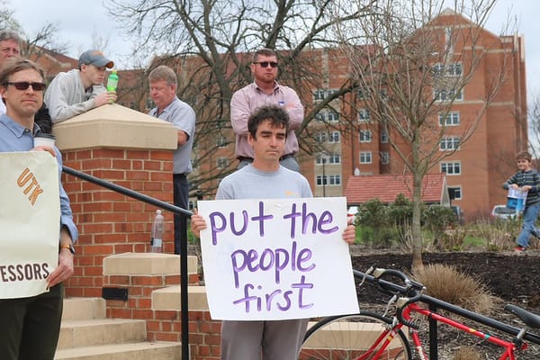 Photo of a group of men protesting against outsourcing. A man in the centre of the frame holds a sign saying "put the people first". 
