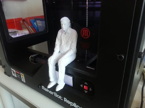 A white, 3D-printed human figure, sitting on the printing platform of a MakerBot Replicant 3D printer.